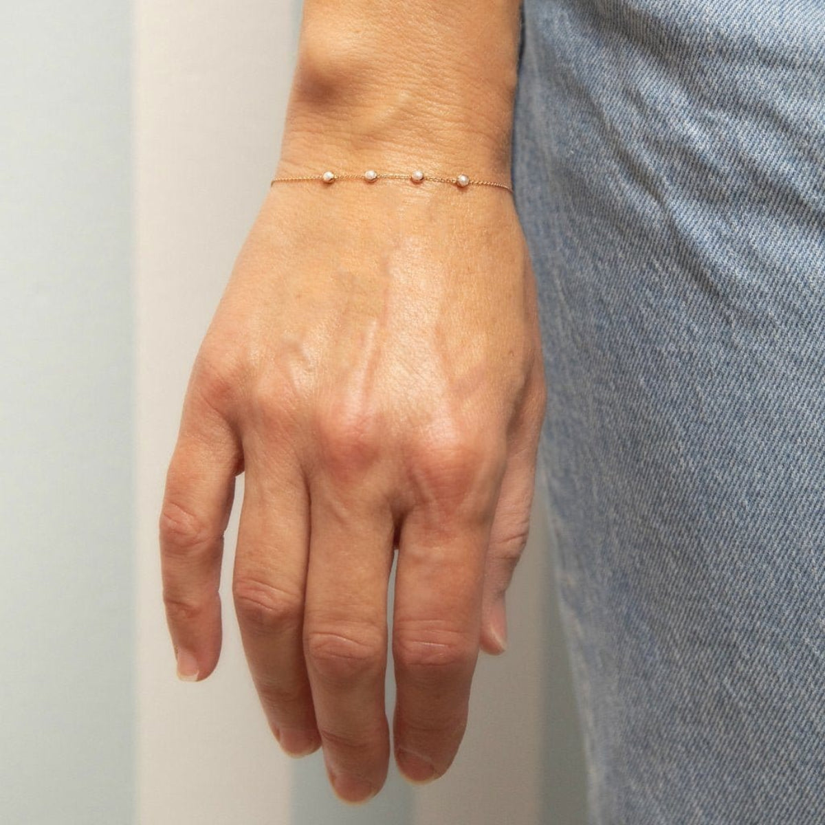 A hand wearing a Custom Gold Bracelet - 4 Letters with four pearl birthstones next to light blue jeans on a neutral stripe background. 