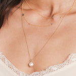 Close-up of a woman's decellotage wearing a 14K Gold Asymmetrical Charm Necklace -Angel Wing, a Custom Gold Necklace - 4 Letter,  and a Perla Pendant on a Classic Bead Chain, with a white lace top. 