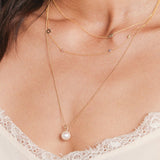 Close-up of a woman's decellotage wearing a 14K Gold Asymmetrical Charm Necklace -Angel Wing, a Custom Gold Necklace - 4 Letter,  and a Perla Pendant on a Classic Bead Chain, with a white lace top. 