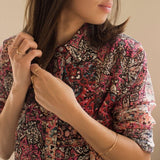 A woman wearing several yellow gold rings, including a Petal Ring, Geo Stacking Ring Triangle and Geo Stacking Ring Square, along with a Flirt Bangle, styled in a colourful patterned top against a light neutral background. 