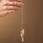 A hand holding three Retreat Necklaces in Clarity, Happiness and Love, a Geo Stacking Ring Square and a Birthstone Ring on a light brown background. 