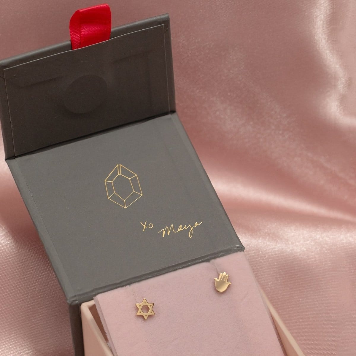 Charm Studs - Star of David and Hamsa in yellow gold in a Maya Brenner jewellery box against a light pink silk background. 