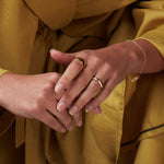 A pair of hands wearing a yellow gold Thick Band, Tube Ring and Love Bracelet, with the person wearing mustard yellow clothing. 