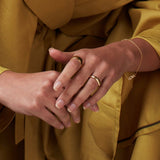A pair of hands wearing a yellow gold Thick Band, Tube Ring and Love Bracelet, with the person wearing mustard yellow clothing. 
