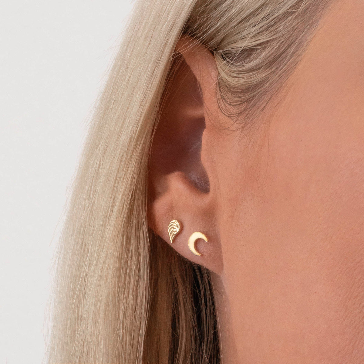 A close up of a yellow gold angle wing stud nd a yellow gold crescent mood stud on a woman's ear