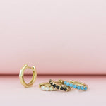 An Alli Webb Faceted Huggie, Opal Huggie, Onyx Huggie and Turquoise Huggie laid on a light pink surface. 