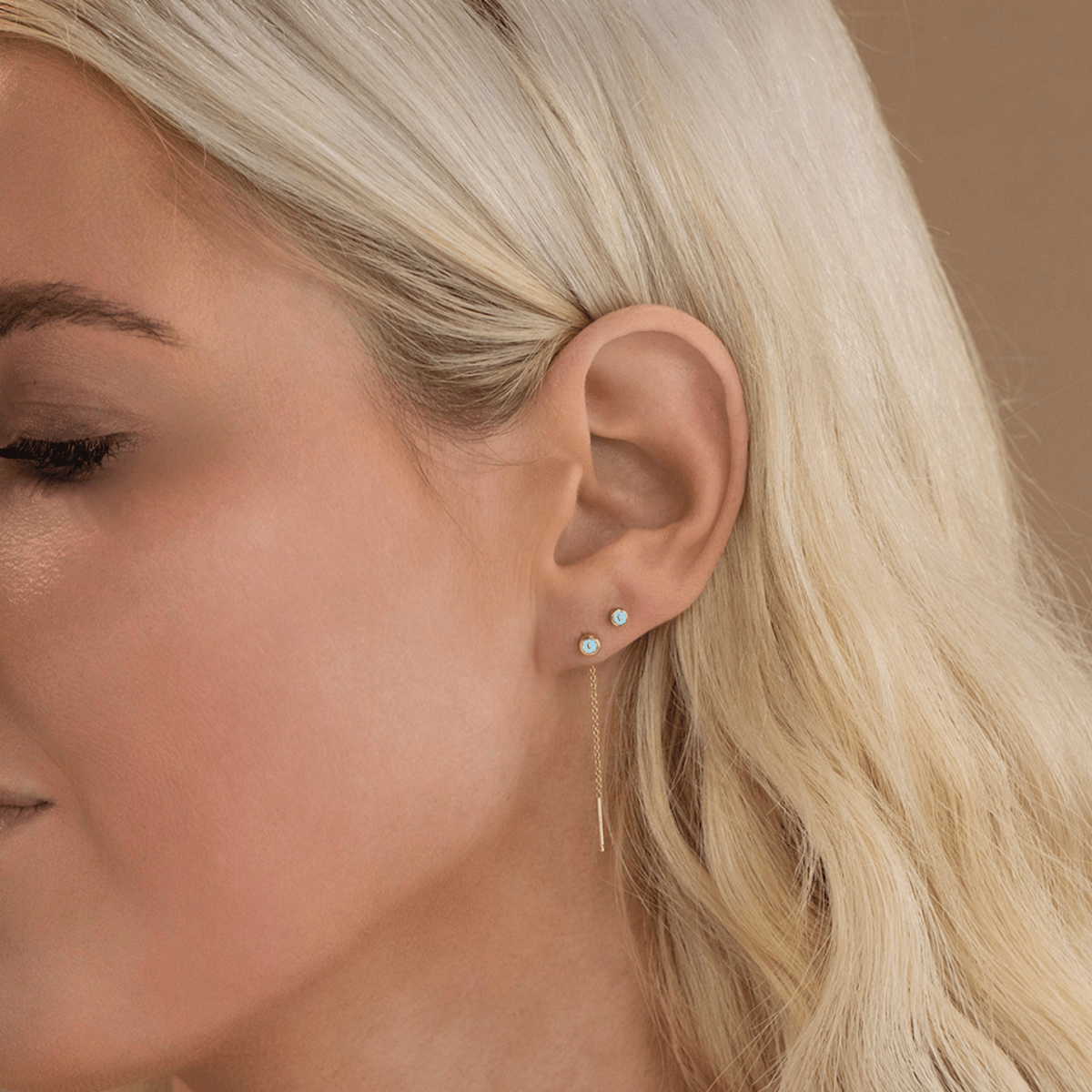 Close-up of a Birthstone Stud and Threader Earring in Aquamarine (March) in a blonde woman's ear against a light brown background. 