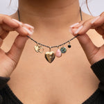 Close-up of a woman's décolletage wearing a Black Rhodium Antique Bead Chain with multiple charms - an Engraved Heart Charm, a Diamond Heart Locket Charm, a Pink Opal Tear Drop Charm, a Small Abalone Cabochon Charm, an Onyx Dangle Charm and a Cut Out Heart Charm, with her hands holding the chain and a black top against a light grey background. 