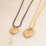 A Black Rhodium Antique Bead Chain with a Heart Locket Charm and a yellow gold Box Chain with a Diamond Heart Locket Charm against a beige marble background. 