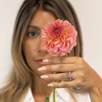 A woman wearing several stacked Bouquet Rings (Periwinkle, Violet, Lily and Peony), styled with a white shirt and holding a pink flower against a softly blurred light background.