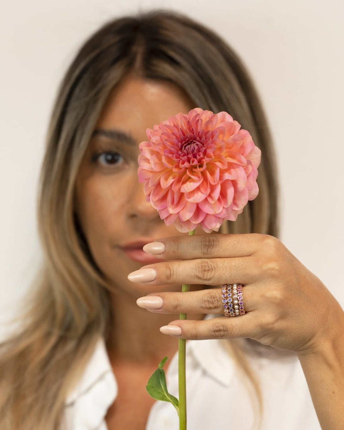 A woman wearing several stacked Bouquet Rings (Periwinkle, Violet, Lily and Peony), styled with a white shirt and holding a pink flower against a softly blurred light background.