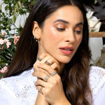 A brunette woman wearing several Bouquet Rings, a Pave Huggie, Gold Safety Pin Earring and a Pave Safety Pin Earring, with a white lace top against a floral background. 