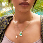 Close-up of a woman wearing a Large Abalone Cabochon Charm on a Box Chain, Perla Necklace and Diamond Cascade Earring, styled in a back and sage green checkered top, with a blurred outdoors background. 