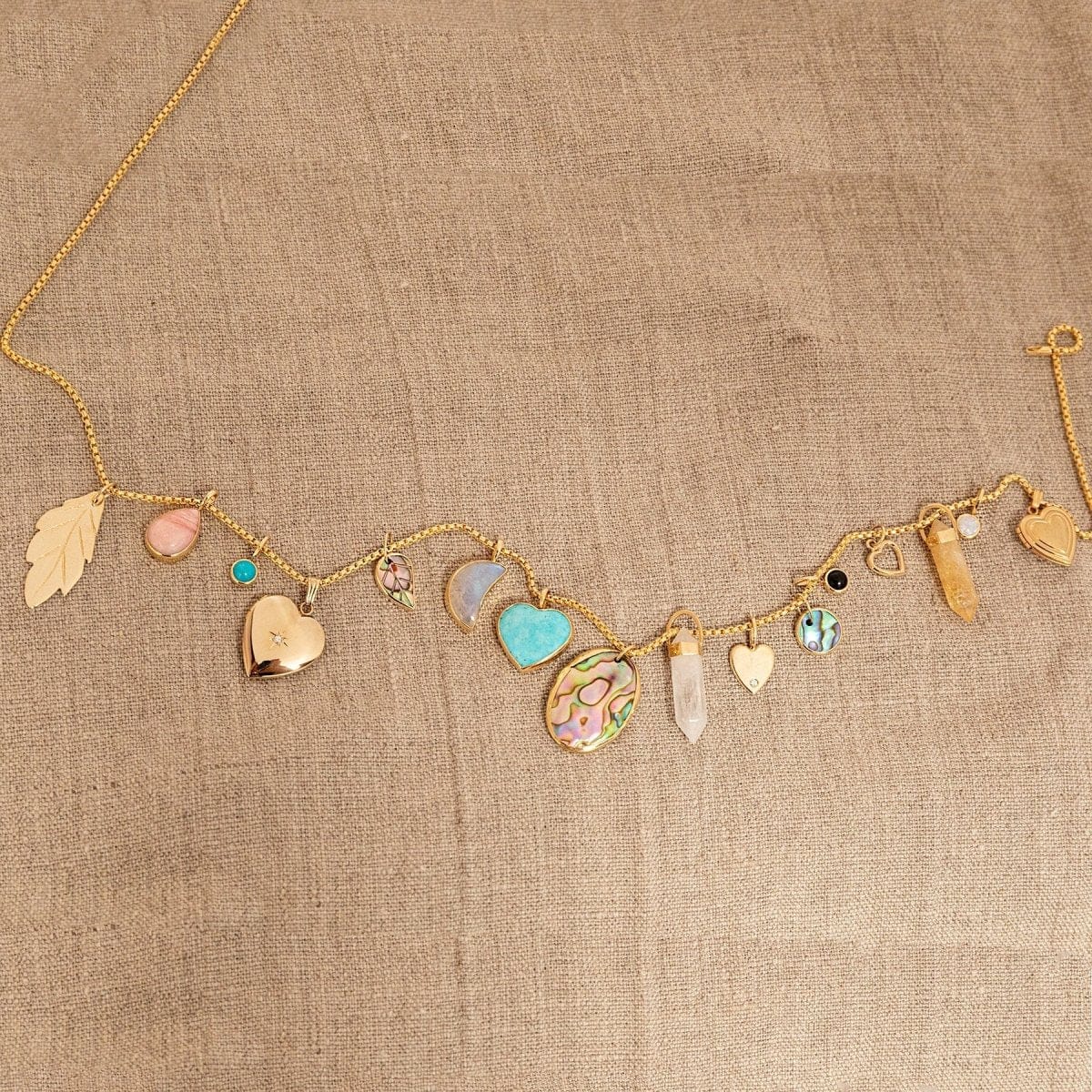 A Box Chain with the following charms - 
20th Anniversary Leaf Pendant, Pink Opal Tear Drop Charm, Turquoise Dangle Charm, Diamond Heart Locket Charm, Abalone Leaf Charm, Moonstone Crescent Charm, Turquoise Heart Charm, Large Abalone Cabochon Charm, two Balance Crystal Charms, Engraved Heart Charm, Small Abalone Cabochon Charm, Onyx Dangle Charm, Cut Out Heart Charm, Opal Dangle Charm and a Heart Locket Charm, against a beige fabric background. 