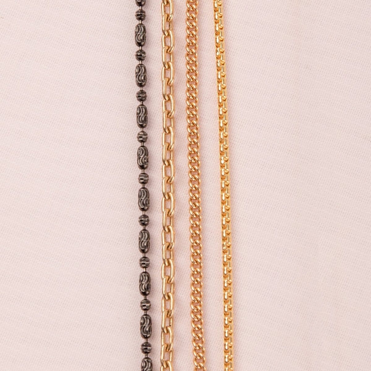 A Black Rhodium Antique Bead Chain, a Round Curb Chain, a Box Chain and a Oval Link Chain laid on an off-white fabric background.