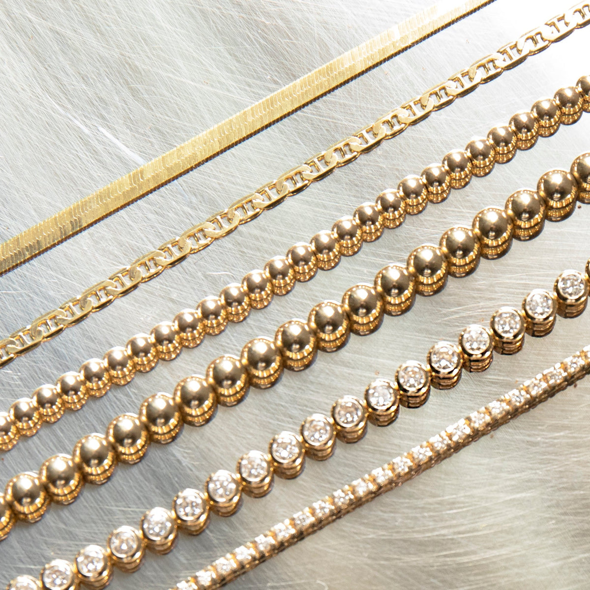 A sequence gold chain next to an overboard gold chain next to two beaded chains next to 2 diamond chains on a stainless steel background.