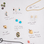 Collection of various jewellery items on white paper with labels, including a yellow Box Chain, Clarity Crystal Charms, Etched Turquoise Heart Charms, Small Abalone Cabochon Charms, Dangle Charms (Turquoise, Onyx and Opal), a Moonstone Crescent Charm, a Heart Locket Charm, a Large Abalone Cabochon Charm, a Pink Opal Tear Drop Charm and a Black Rhodium Antique Bead Chain.