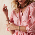 A woman wearing multiple coloured Bouquet Necklaces, Bouquet Bracelets, yellow gold Bubble Bracelets and rings, styled in a pink dress. 