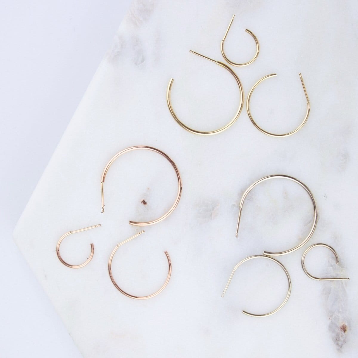 Crescent Hoops / Small, Medium and Large in rose, white and yellow gold on a white marble surface. 