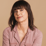 A woman wearing a Custom Mixed Gold Necklace with two birthstones and a hamsa charm, styled in a pink shirt against a beige background. 