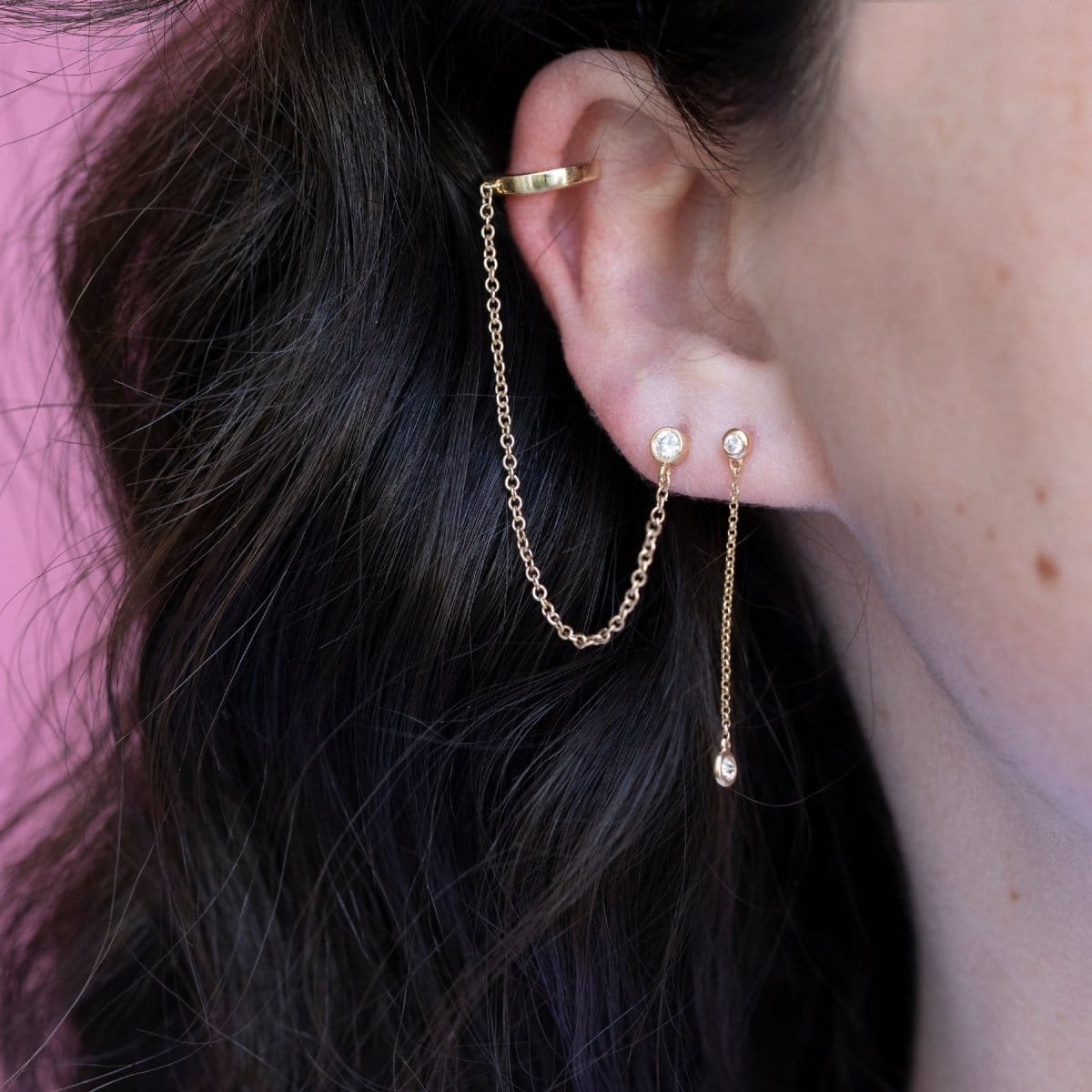 Close-up of a Coupling Ear Cuff + Stud and Droplet Diamond Earrings in yellow gold in a brunette woman's ear against a pink background. 