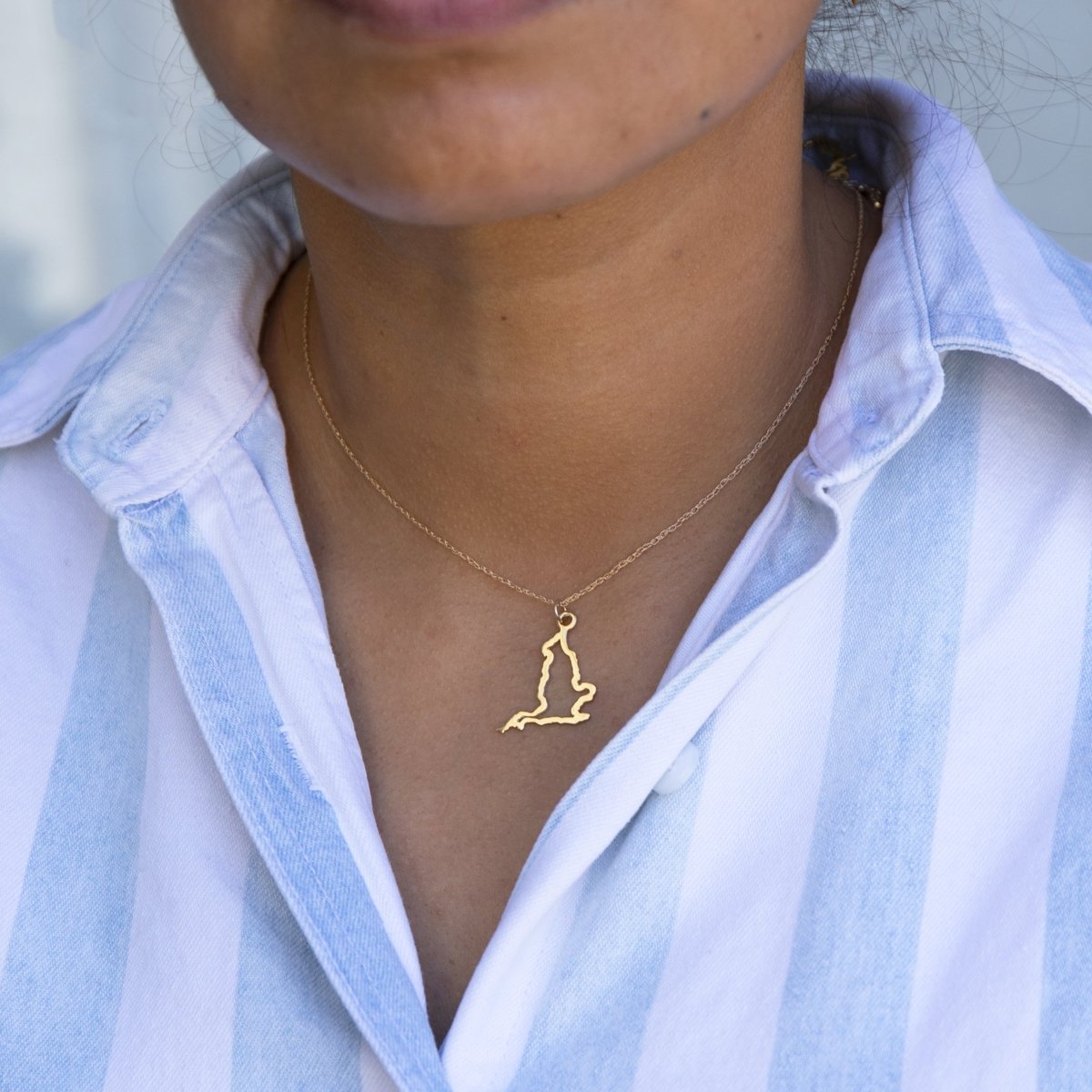 Close-up of a person's neck wearing an England necklace in yellow gold with a blue and white striped shirt against a light grey blurred background. 