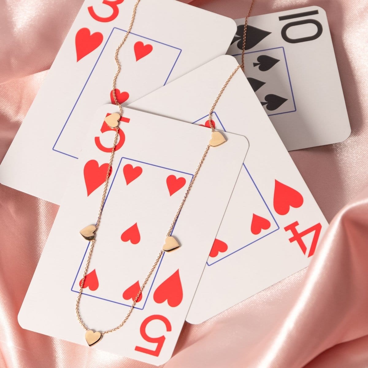 A rose gold 5 of Hearts Necklace on top of playing cards and a pink silk fabric background. 