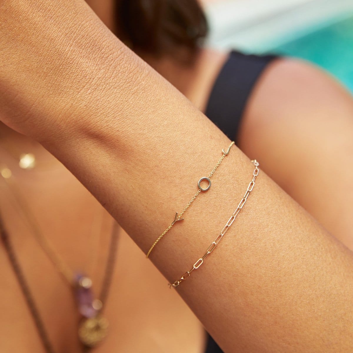 A wrist wearing an Element Short Link Bracelet and a Custom Gold Bracelet with 3 letters (J, O and Y), with a blurred background.
