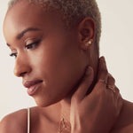 A woman with her hand on her neck, wearing three earrings – a pavé letter M stud, a threader earring with a gold heart stud and a gold letter B stud, along with various rings and necklaces.
