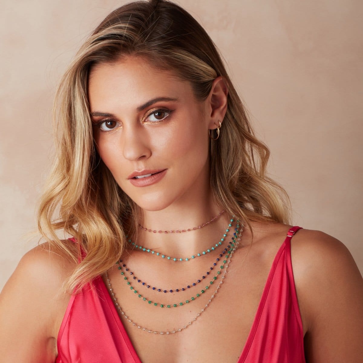 A blonde woman wearing several Bouquet Necklaces (Peony, Bluebell, Thistle, Clover and Sage), yellow gold hoop earrings and a dark pink top against a neutral background. 