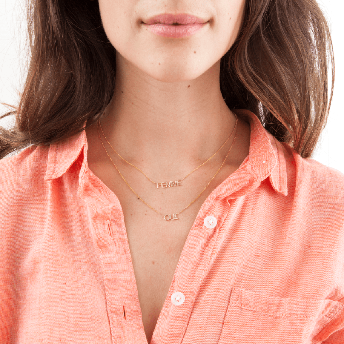 A close-up of a brunette woman wearing a FEMME Necklace and an OUI Necklace, with a light orange shirt against a white background.
