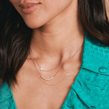 A close-up of a woman's neck wearing two 14k Gold Asymmetrical Charm Necklaces with star and moon charms, along with a Diamond Arc Layering Necklace in rose or white gold, styled in a turquoise blue shirt.
