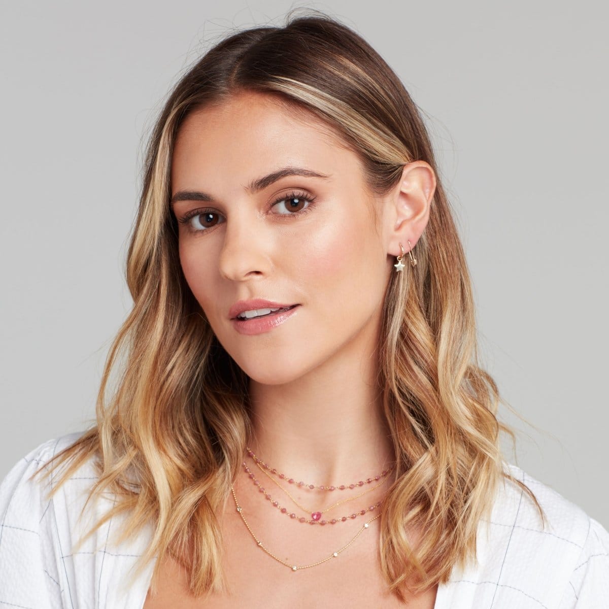 A blonde woman wearing The Peony Bouquet Necklaces, The Sweetheart Necklace - Pink Sapphire, Quintet Diamond Layering Necklace, Charmed I'm Sure Hoop / Star and a Gold Safety Pin Earring, with a white linen top and a light grey background. 