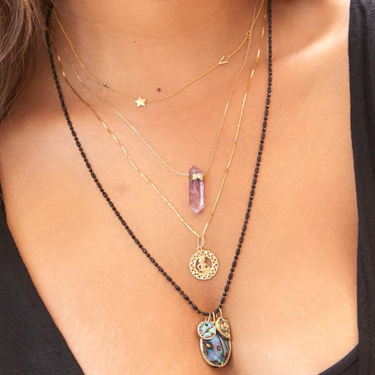 A close-up of a woman's décolletage wearing a yellow gold Custom Gold Necklace - 4 Letters, The Healing Retreat Necklace, a Zodiac Necklace - Gemini and  a Black Rhodium Antique Bead Chain with a Large Abalone Cabochon Charm, Abalone Leaf Charm and a Small Abalone Cabochon Charm, with a black top. 
