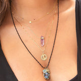 A close-up of a woman's décolletage wearing a yellow gold Custom Gold Necklace - 4 Letters, The Healing Retreat Necklace, a Zodiac Necklace - Gemini and  a Black Rhodium Antique Bead Chain with a Large Abalone Cabochon Charm, Abalone Leaf Charm and a Small Abalone Cabochon Charm, with a black top. 