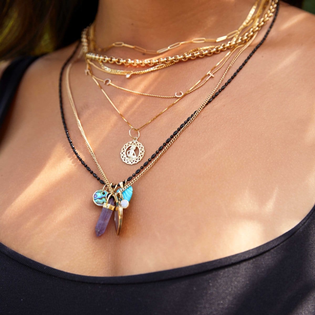 Close-up of a woman's décolletage wearing an Element Long Link Chain, Rolo Necklace, Overboard Layering Necklace, Custom Gold Necklace – 4 Letters, Single Diamond Layering Necklace, Zodiac Necklace – Gemini, a Black Rhodium Antique Bead Chain with an Etched Turquoise Heart Charm, Small Abalone Cabochon Charm and Onyx Dangle Charm, and a Box Chain with a Balance Crystal Charm, Diamond Heart Locket Charm and Opal Dangle Charm, wearing a black tank top.