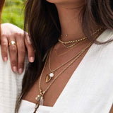 Close-up of a woman's décolletage wearing a Rolo Necklace, Birthstone Arc Layering Necklace, a Monogram Necklace with Diamond, a Round Curb Chain with a Diamond Heart Locket Charm and a Perla Pendant, an Oval Link Chain with a Moonstone Crescent Charm and a Balance Crystal Charm, and a Thick Band, 
all in yellow gold, with a white top with a green blurred background. 