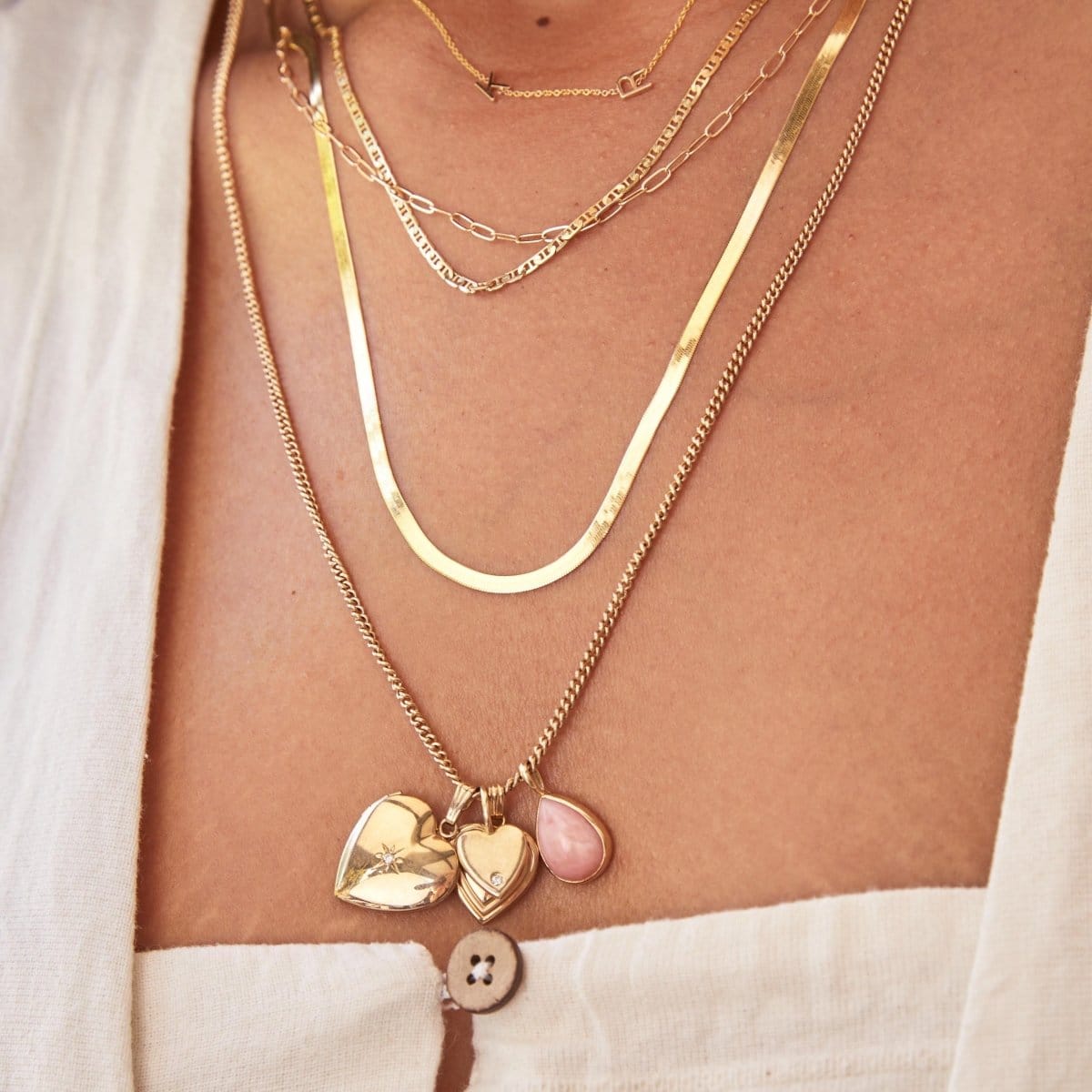 Close-up of a woman's decellotage  area wearing a Custom Gold Necklace – 2 Letters, Overboard Layering Chain, Element Short Link Chain, Sequence Chain and Round Curb Chain with a Pink Opal Tear Drop Charm, Heart Locket Charm, Diamond Heart Locket Charm and Engraved Heart Charm, styled in a white linen top. 
