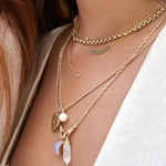 Close-up of a décolletage wearing a Rolo Necklace, a Diamond Layering Necklace, a Birthstone Arc Layering Necklace, a Monogram Necklace with Diamond, a Round Curb Chain with a Diamond Heart Locket Charm and a Perla Pendant, an Oval Link Chain with a Moonstone Crescent Charm and a Balance Crystal Charm, all in yellow gold, with a white top and some orange hair visible. 