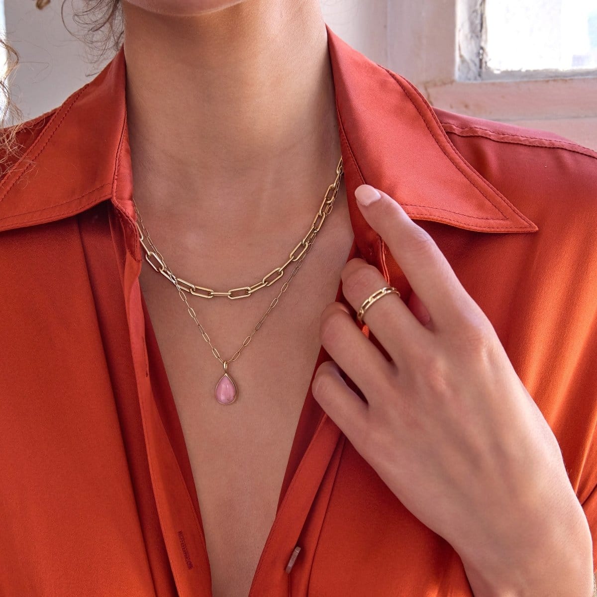A close-up of a woman's décolletage wearing a yellow gold Bold Link Chain, an Element Short Link Chain with a Pink Opal Tear Drop Charm and a Link Ring, with an orange shirt against an indoor background.