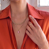 A close-up of a woman's décolletage wearing a yellow gold Bold Link Chain, an Element Short Link Chain with a Pink Opal Tear Drop Charm and a Link Ring, with an orange shirt against an indoor background.