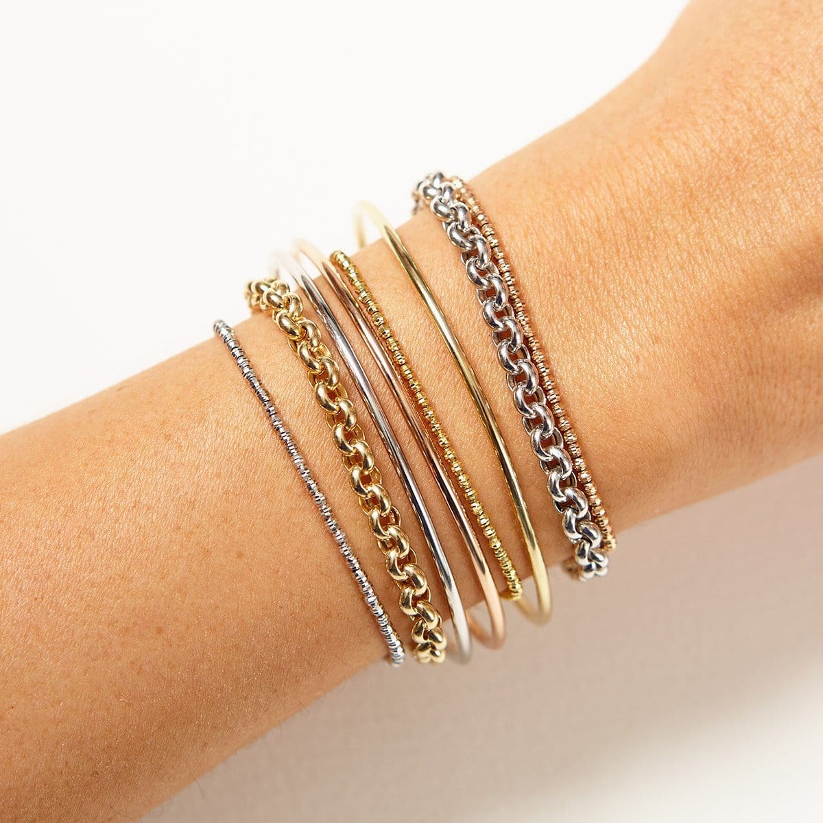 A close-up of an arm wearing mixed metal bracelets: two Rolo Bracelets, three Diamond Cut Bracelets and three Simple Bangles against a white background.