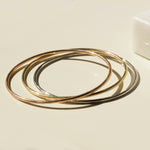 Three Simple Bangles, one in yellow gold, one in rose gold and one in white gold, stacked on a light beige surface. 