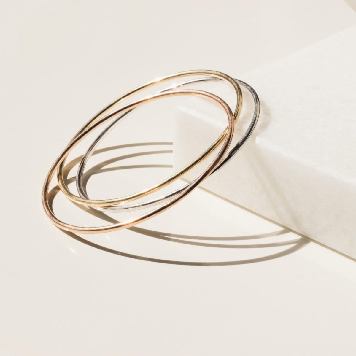 Three Simple Bangles, one in yellow gold, one in rose gold and one in white gold, against an off-white background.