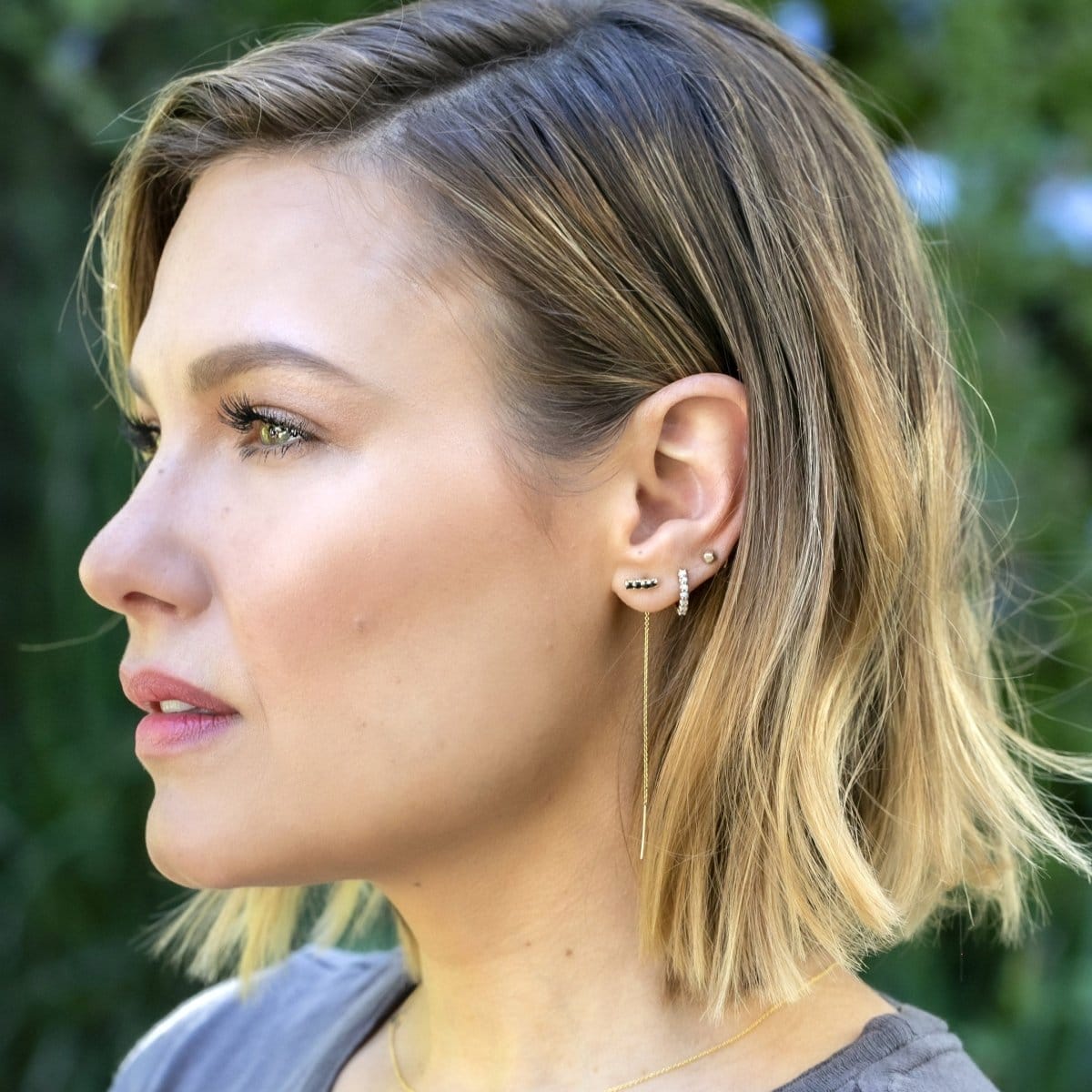 A woman wearing an Alli Webb Onyx Threader, Opal Huggie and a Faceted Comfort Stud, with blonde hair and a grey top, against a green outdoors background. 