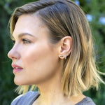 A woman wearing an Alli Webb Onyx Threader, Opal Huggie and a Faceted Comfort Stud, with blonde hair and a grey top, against a green outdoors background. 