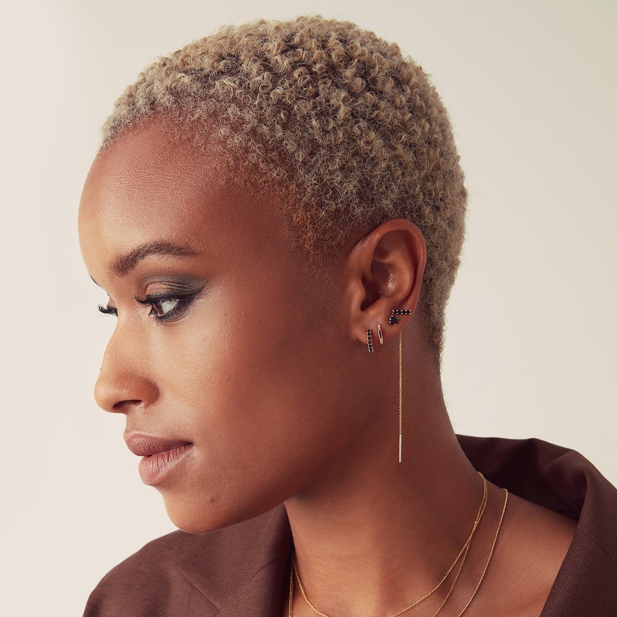 A woman wearing an Alli Webb Onyx Comfort Stud, an Onyx Threader, an Onyx Huggie and a Faceted Huggie, with short blonde hair and a brown top against a neutral background. 