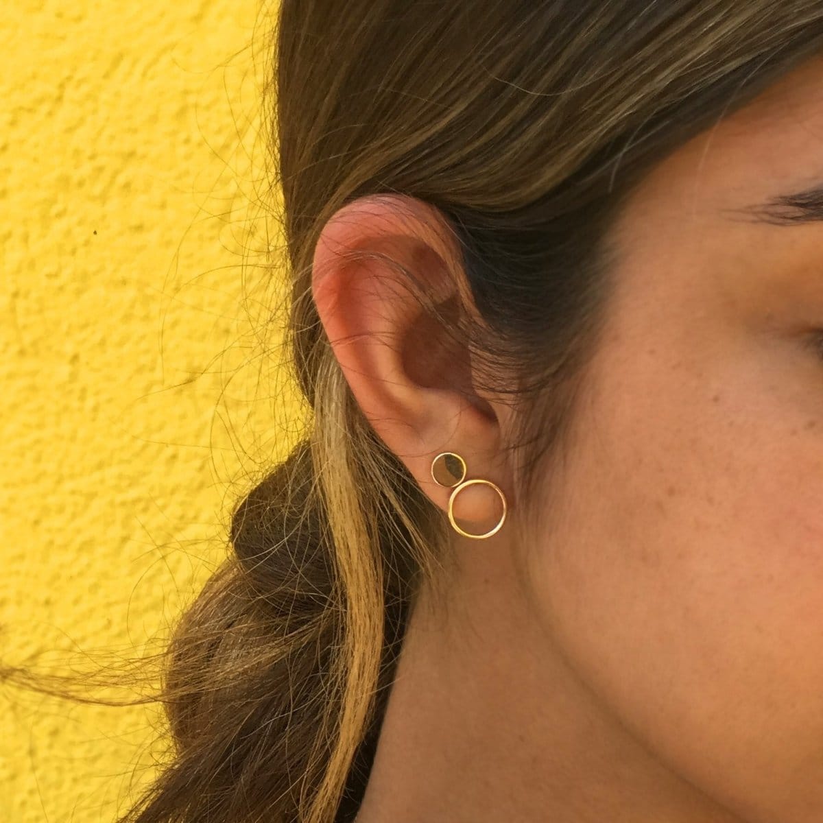 Close-up of an Open Eclipse Earring - Small and Solid Eclipse Earring - Mini in a brunette woman's ear against a yellow background. 