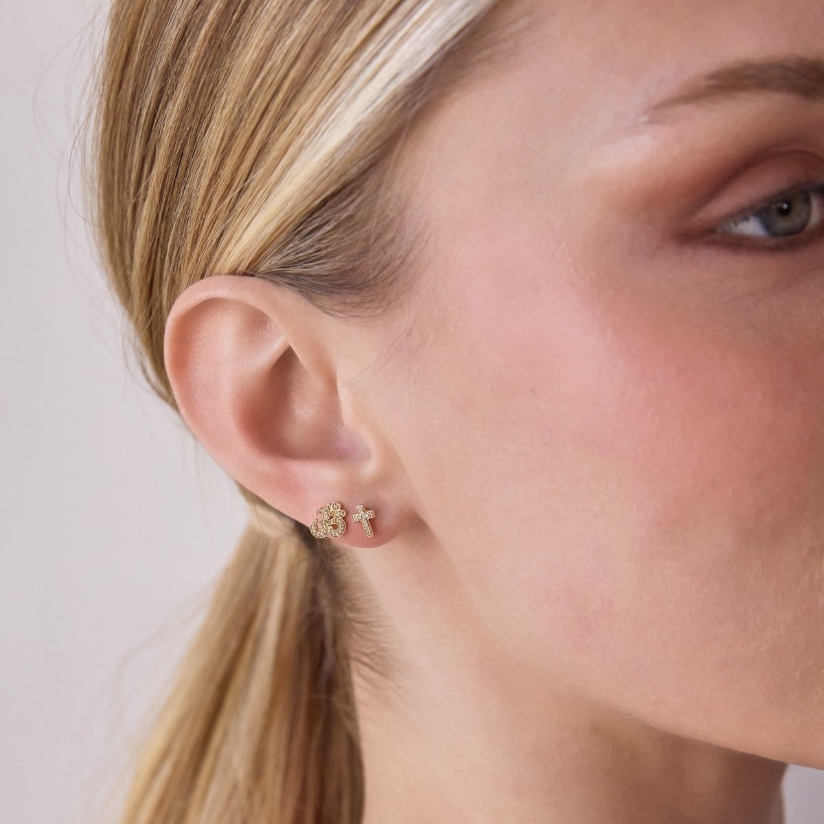 Close-up of Pavé Charm Studs - Cross and Om in yellow gold in a blonde woman's ear against a light grey background. 