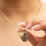 A neck wearing a Round Curb Chain with a Large Abalone Cabochon Charm, Heart Locket Charm and an Opal Dangle Charm, being held by a hand. 